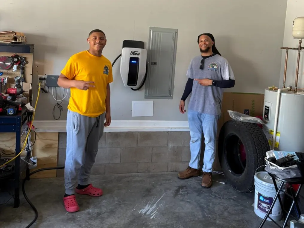 Two Sonshines team members next to a home EV charger and electrical panel in a garage